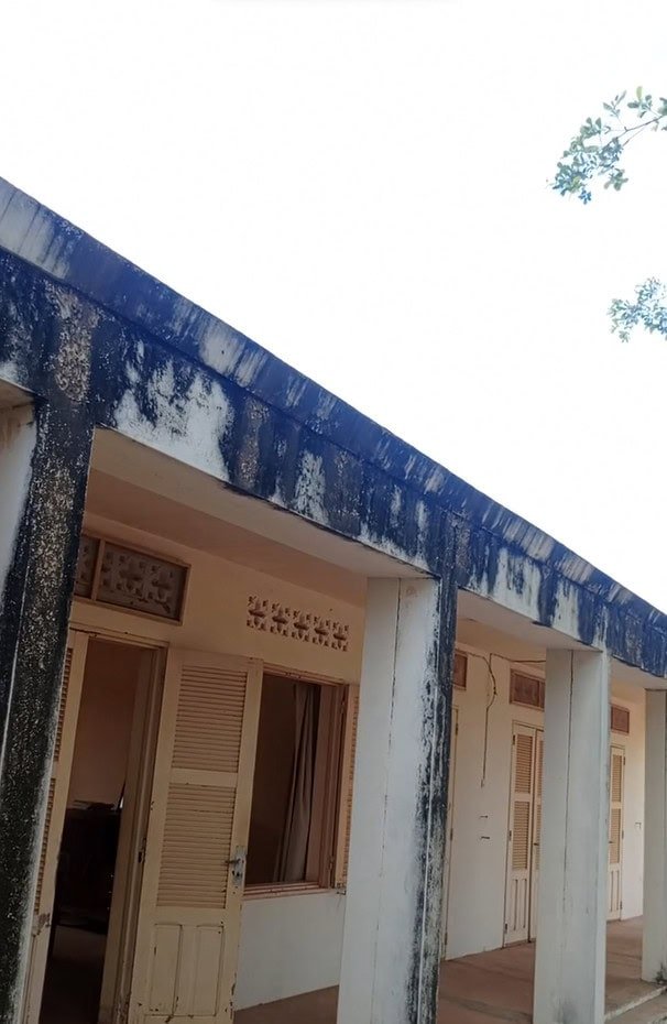 Vieille bâtisse aux murs blancs tachés, aux traces de moisissures bleues et aux portes en bois ouvertes sous un ciel radieux.