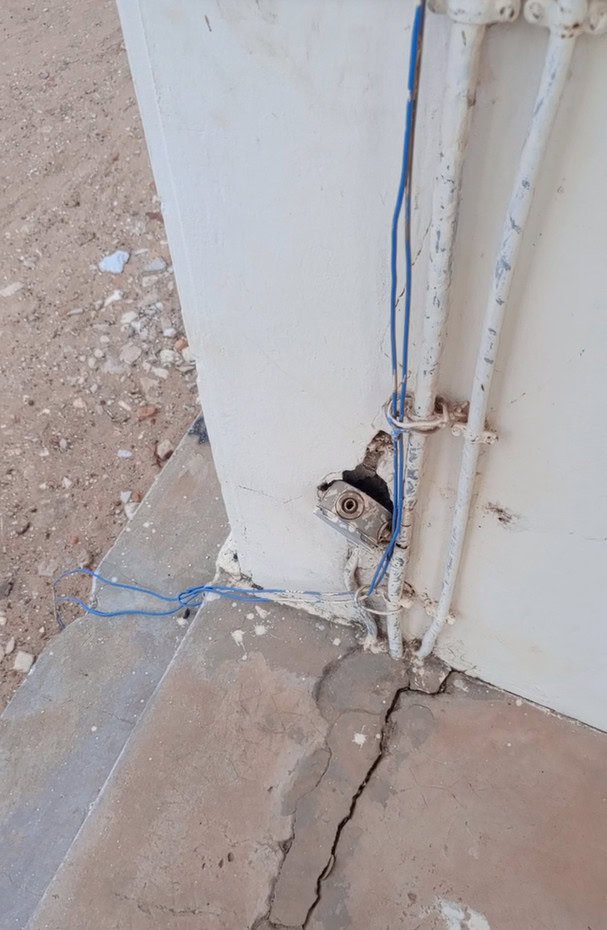 Des fils bleus sortent d'un trou dans un mur blanc près d'un sol en béton fissuré.