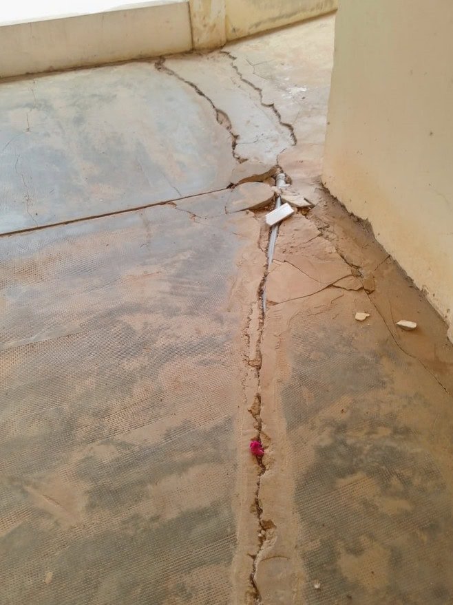 Grande fissure traversant un sol en béton poussiéreux, avec quelques débris et un petit objet rose près de la fissure.