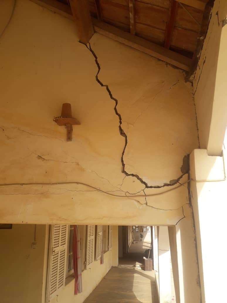 Grande fissure traversant un mur jaune sous un toit de tuiles, avec lumière du soleil et câblage visible.
