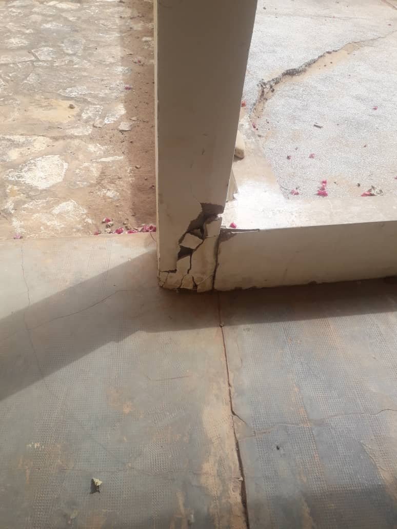 Une colonne en béton avec une grande fissure à la base près d'une zone carrelée et extérieure.